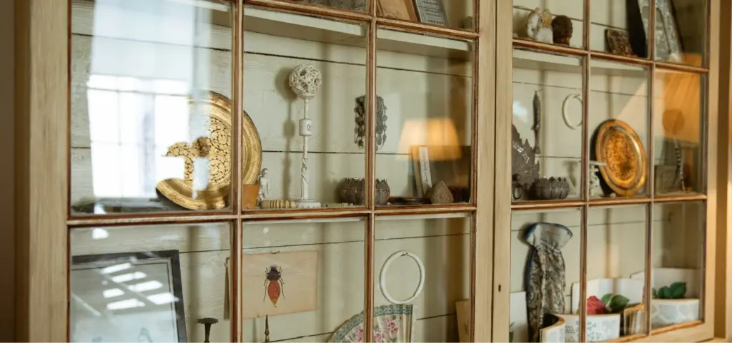 Glass cabinet with a wooden frame showcasing a variety of objects, including decorative plates, a framed insect picture, and small artifacts, all arranged on wooden shelves.