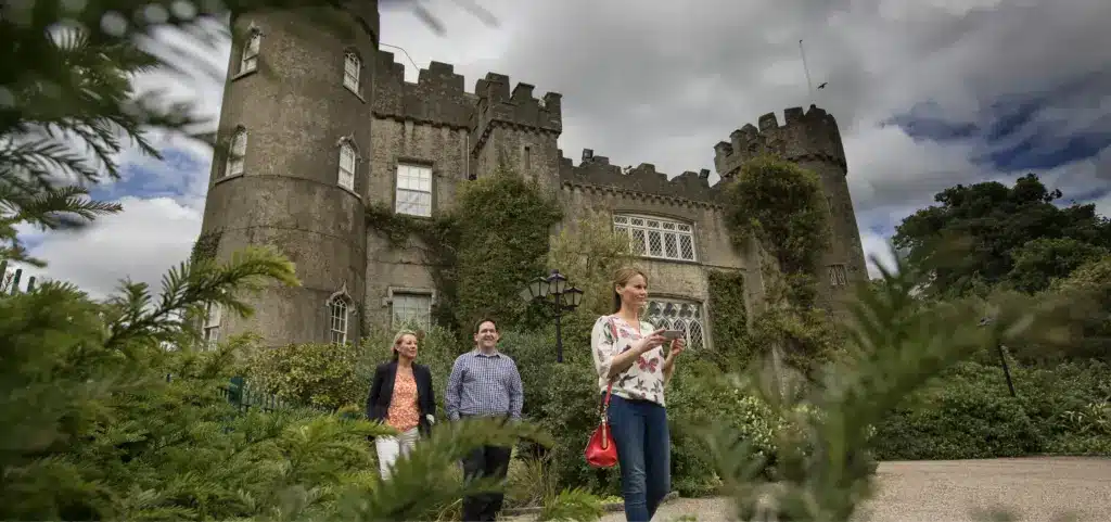 Three people stroll near the ivy-covered stone walls and large windows of Malahide Castle. The sky is cloudy, enhancing the historic charm, and lush greenery envelops the scene. One person is looking at a phone while the others smile warmly.