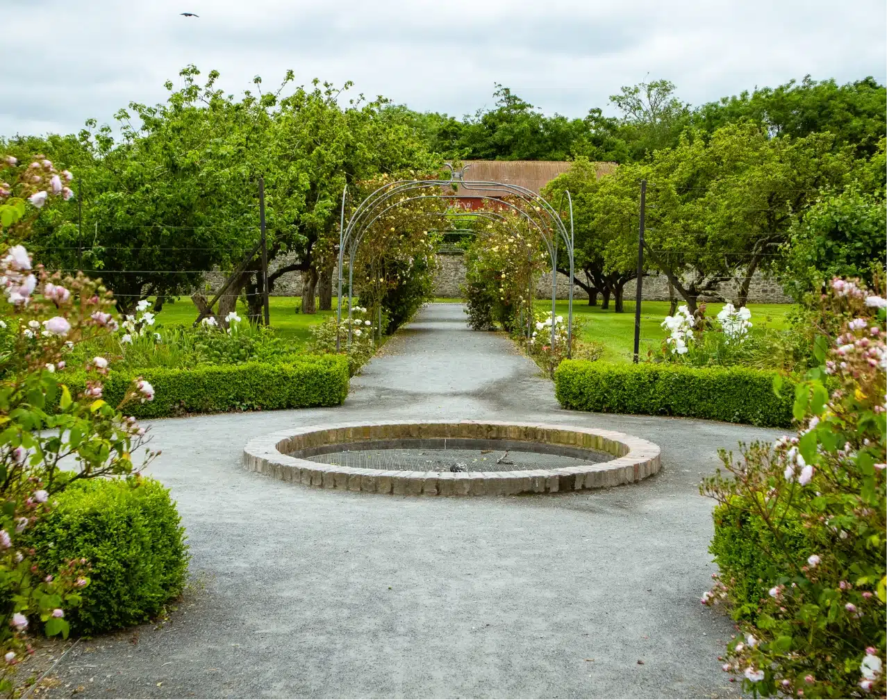 A tranquil garden scene near Malahide Castle features a stone path leading to an archway covered in climbing plants. The path is lined with flower beds and bushes, and a circular stone fountain sits in the center, surrounded by lush greenery and trees.