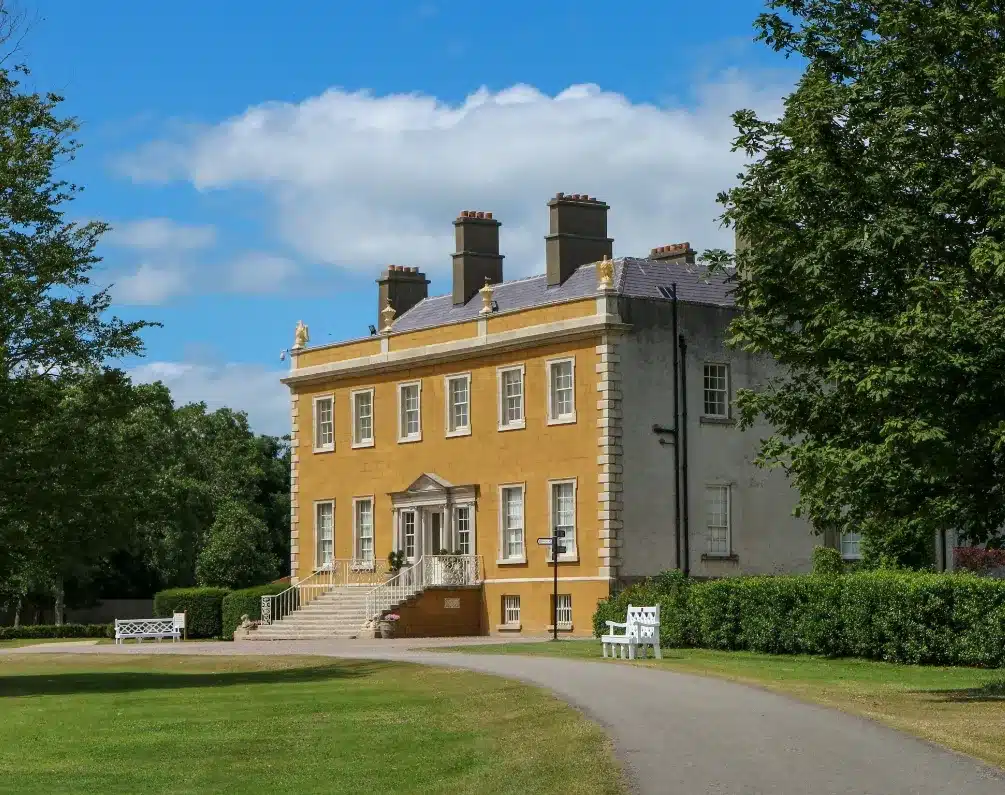 A stately yellow and gray mansion with chimneys stands under a blue sky with scattered clouds. A curved driveway leads to the entrance, flanked by lush green lawns and trees. Benches are placed near the front of the house.