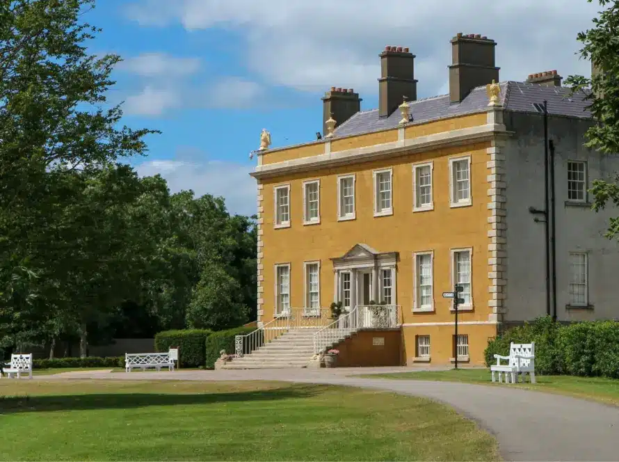 A grand, two-story, mustard-colored mansion reminiscent of Malahide Castle stands behind a curved driveway. Tall chimneys rise above lush green lawns, benches, and trees under a partly cloudy sky.
