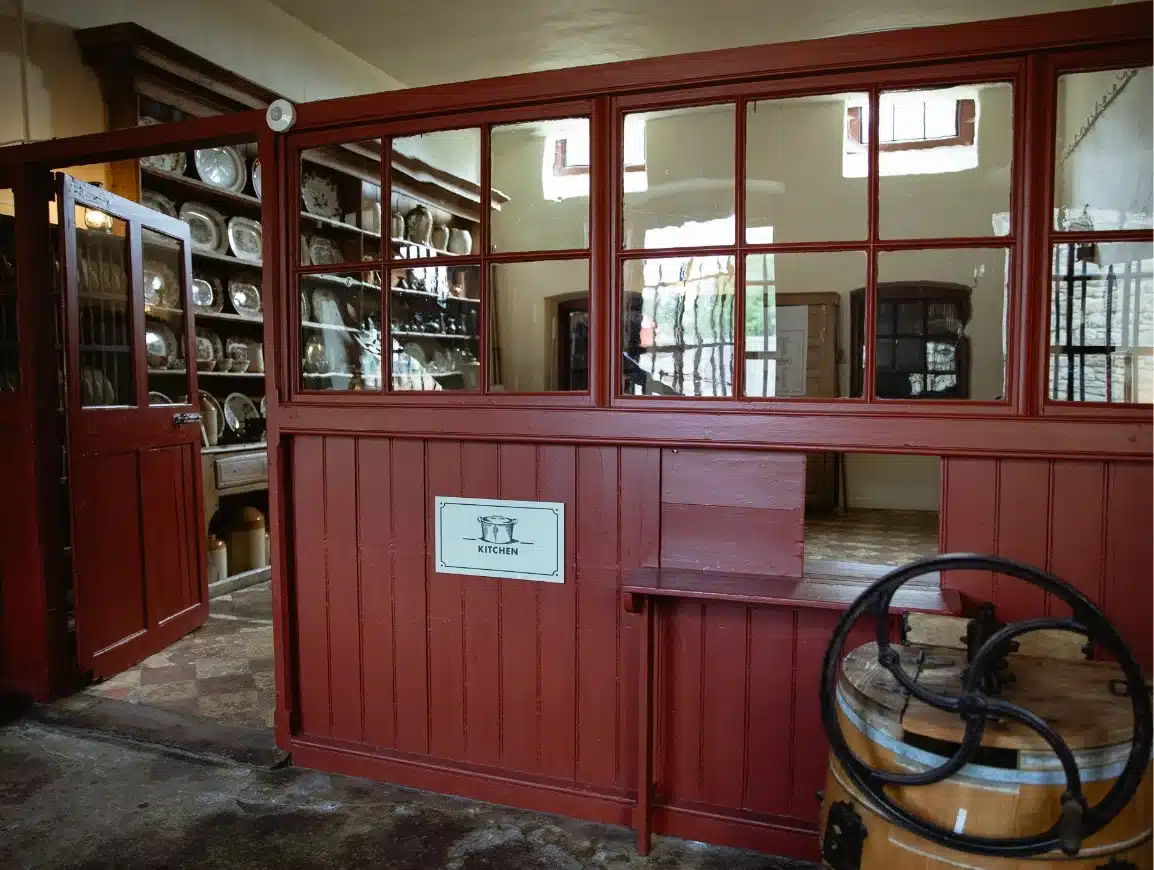 Historic kitchen pantry with red wooden paneling and glass windows. Shelves display various plates and dishes. A sign labeled "Kitchen" is visible on the panels. A wooden barrel with metal bands is positioned nearby.