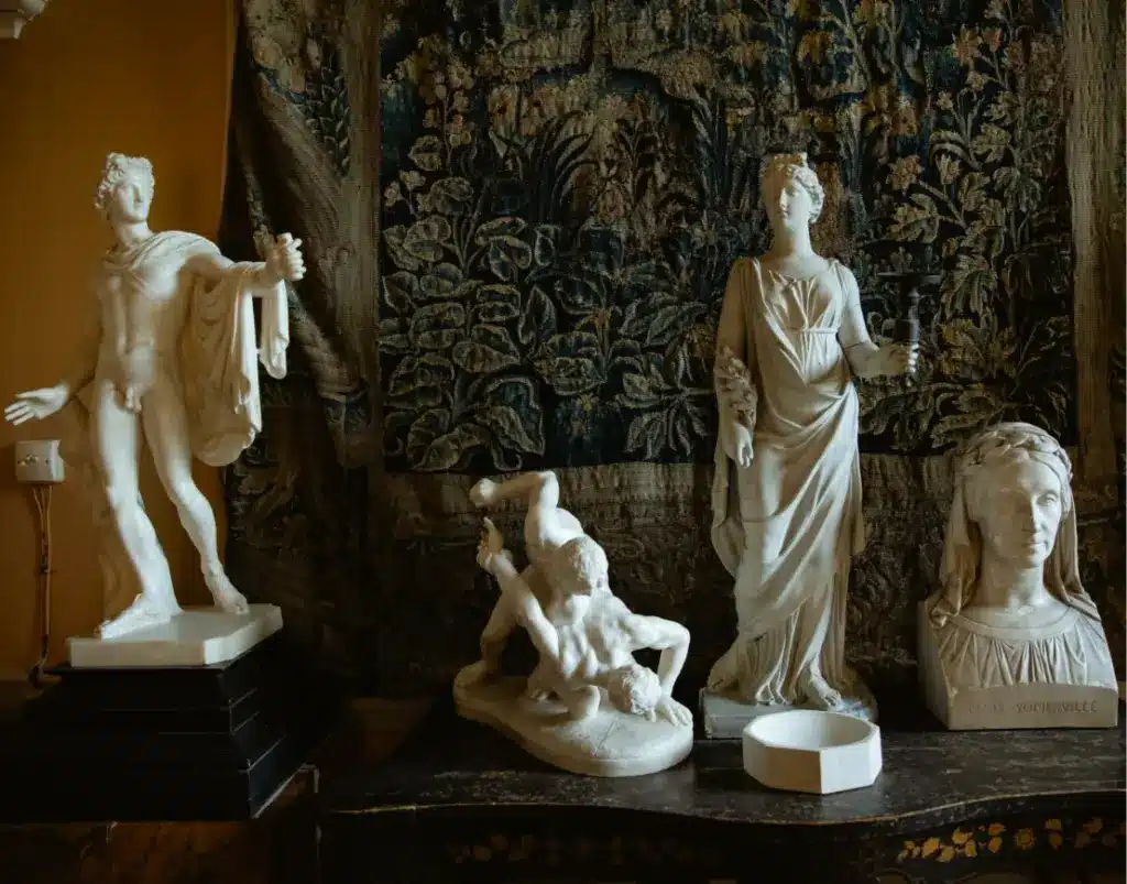 A collection of classical white marble statues on display indoors. The figures include a toga-draped male figure, a dynamic wrestling scene, a standing woman in flowing robes, and a bust of a woman. A detailed tapestry hangs in the background.