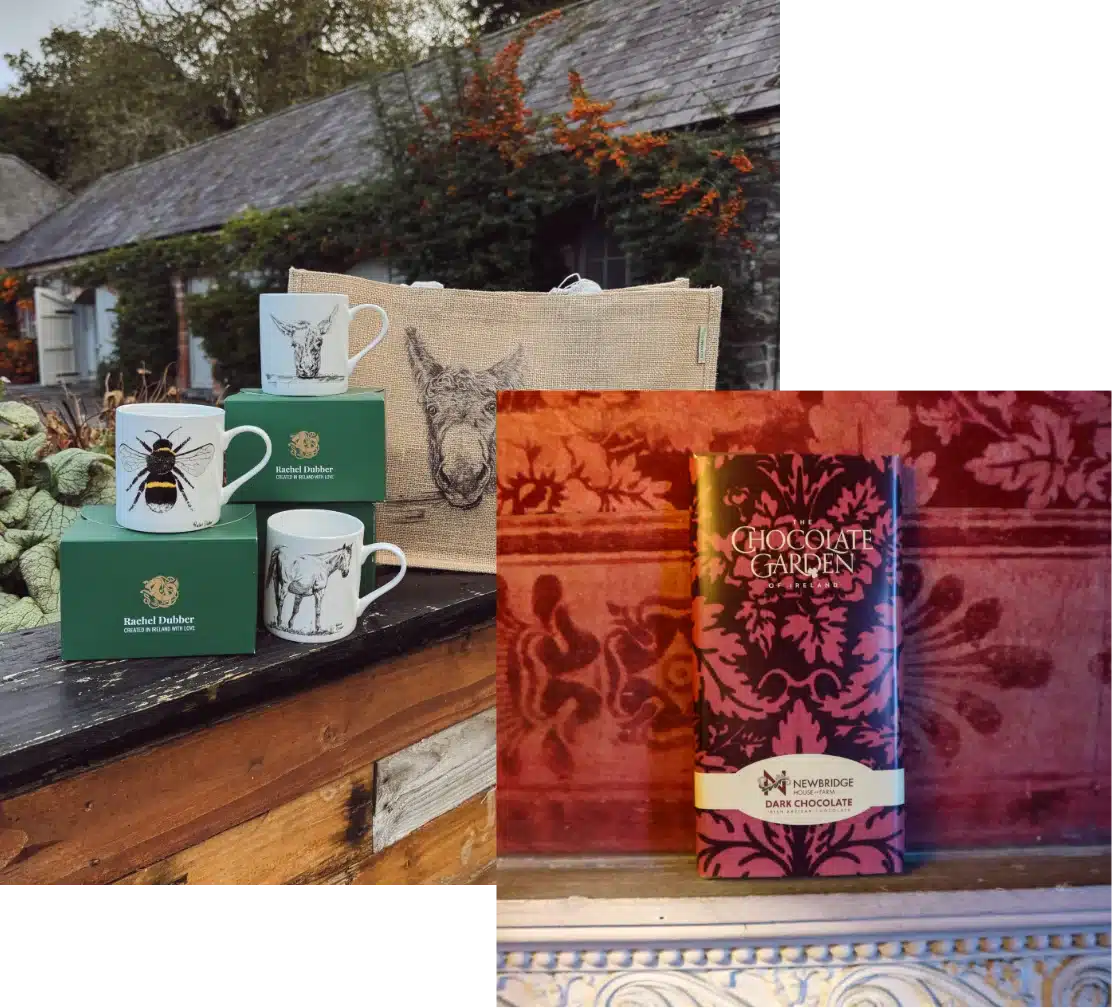 Two mugs adorned with animal illustrations and a donkey-themed tote bag sit on a wooden surface, surrounded by greenery near Malahide Castle. A chocolate bar with a decorative wrapper completes this charming outdoor scene by the rustic building.