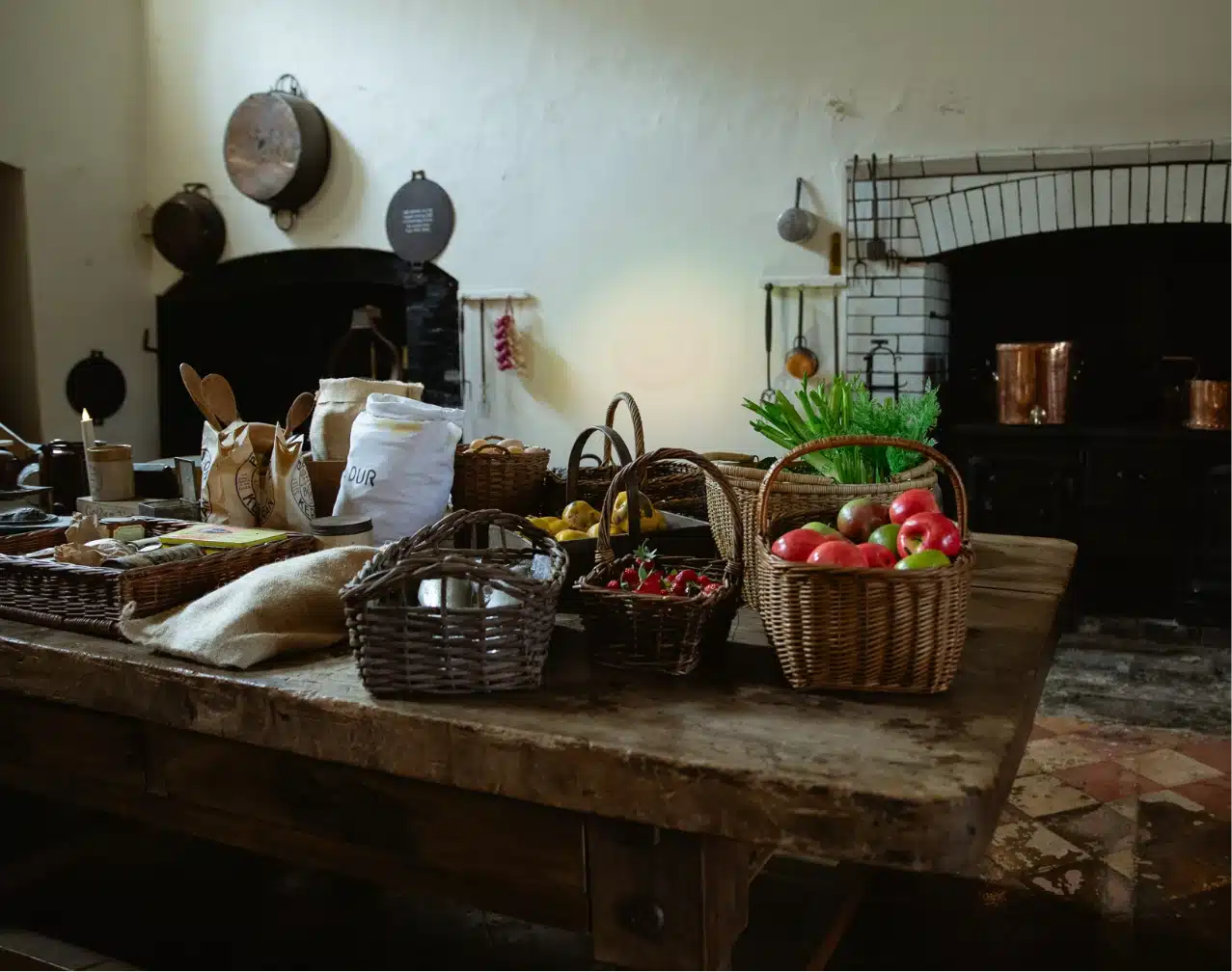 A rustic kitchen reminiscent of Malahide Castle, with a large wooden table covered in wicker baskets filled with apples, chili peppers, and greens. Copper pots and pans adorn the shelves against stone walls, enhancing the vintage ambiance reminiscent of the historic castle's charm.