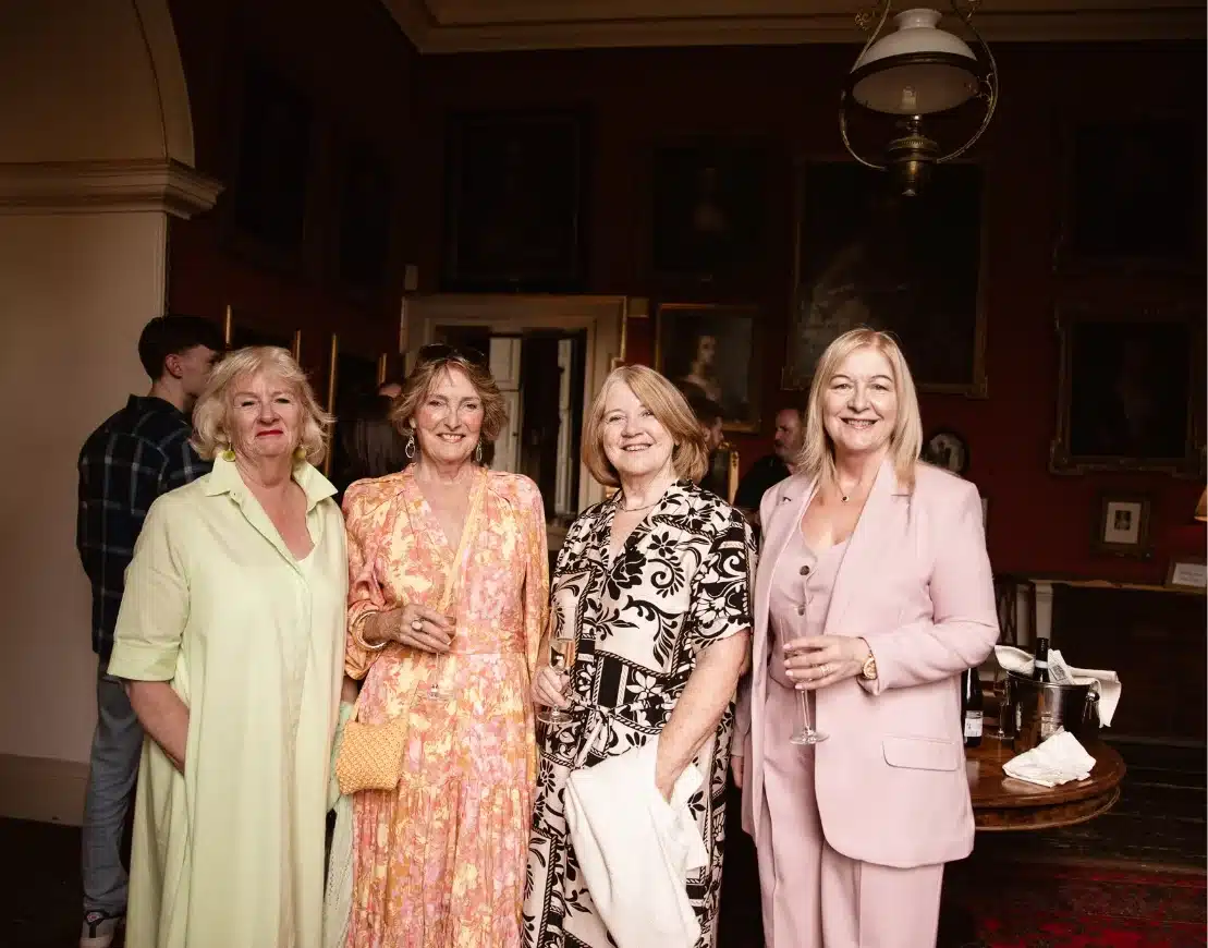 Four women smile and pose for a group photo in a warmly lit room. Three hold drinks, and they wear light-colored, elegant outfits. The background features dark walls with framed paintings and other guests in conversation.