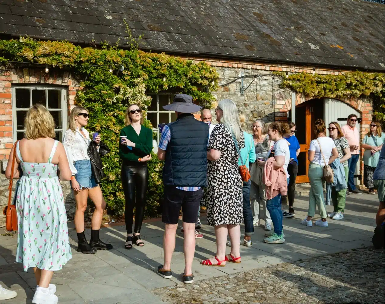 A group of people gathered outside a rustic brick building with ivy, reminiscent of Malahide Castle. Casually dressed, they socialize under sunny skies in a relaxed and friendly atmosphere, some sporting sunglasses.