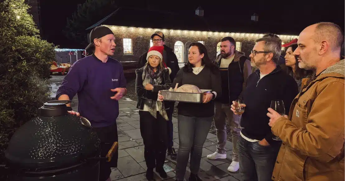A group of people stands outside on a patio at night, reminiscent of gatherings at Malahide Castle. One person is speaking near a green grill, while another holds a tray with a turkey. Some are holding drinks as festive lights adorn the building in the background.