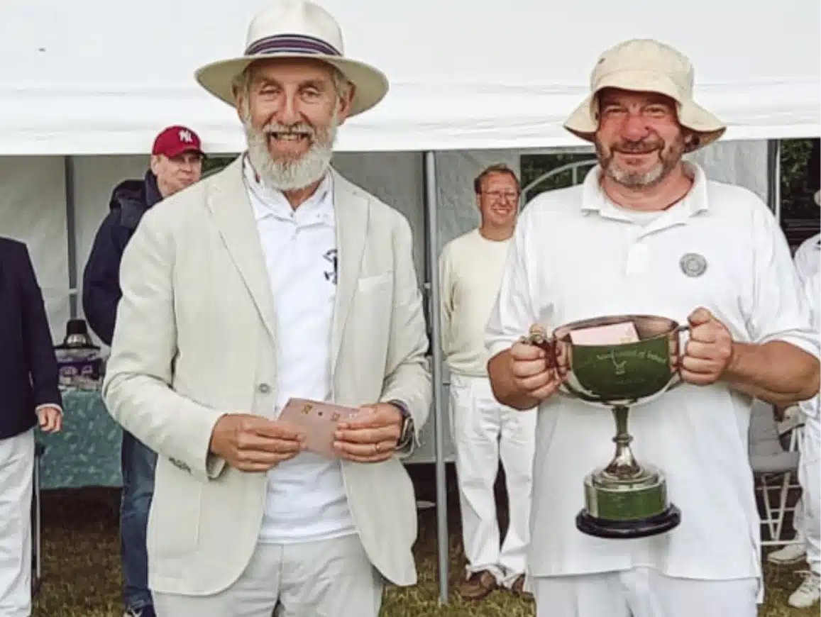 Two men in white attire stand outdoors in front of a tent. One wears a bowler hat and holds a card; the other, wearing a bucket hat, holds a trophy. People in similar attire are in the background.