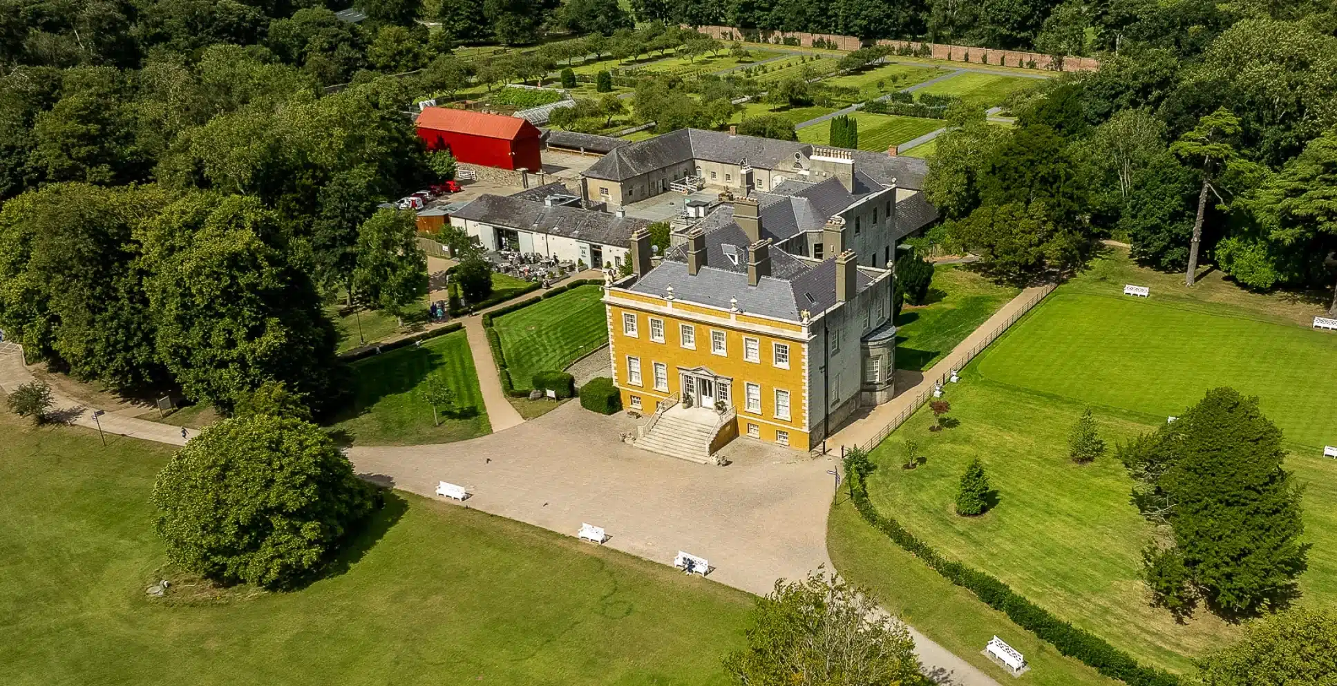 Aerial view of an elegant historic mansion reminiscent of Malahide Castle, with a yellow brick facade, surrounded by manicured lawns, lush gardens, trees, footpaths, and several outbuildings including a large red barn.