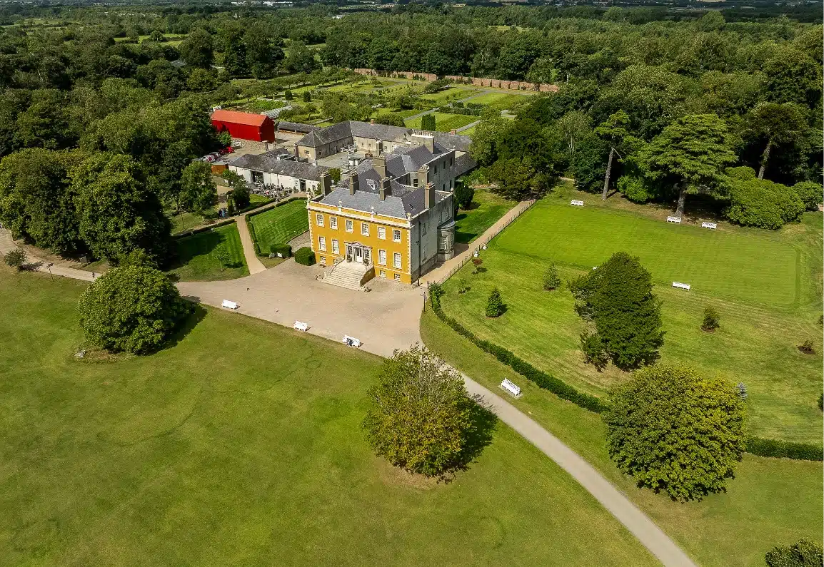 Aerial view of a large, yellow manor house surrounded by green lawns and gardens. The estate includes several outbuildings, pathways, and rows of trees, set in a lush, wooded landscape under a clear sky.