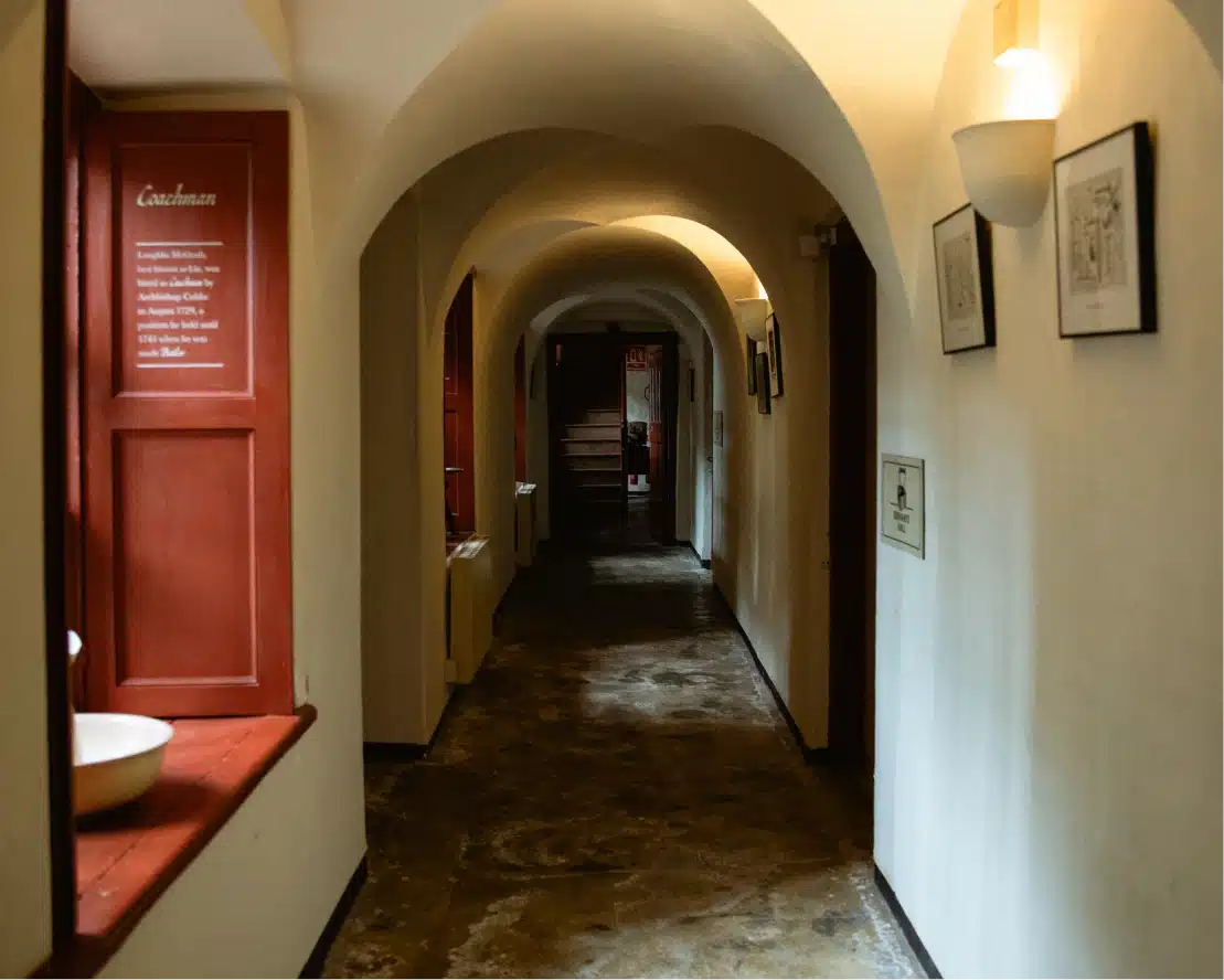 A dimly lit corridor with arched ceilings and a stone floor evokes the historic charm of Malahide Castle. A red-framed window on the left has text beside it, while framed pictures hang on the right wall. At the far end of this enchanting hallway, stairs beckon exploration.