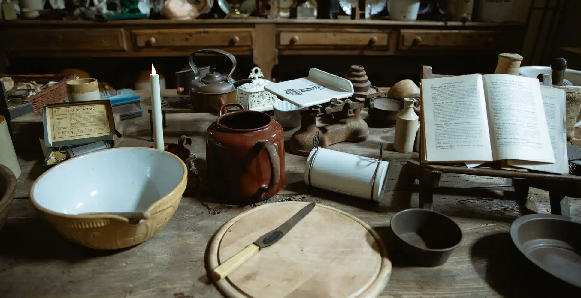 A rustic kitchen table with vintage utensils, an open cookbook, a cutting board with a knife, and a ceramic bowl evokes the timeless charm of Malahide Castle, with a brown teapot, lit candle, and old-fashioned kitchen items scattered around.