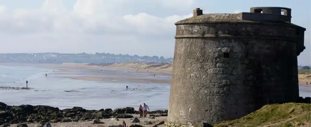 A historic stone tower stands on a rocky coastline with a sandy beach stretching into the distance. People are walking along the beach, and the sky is partly cloudy. The sea is calm, and a distant town is visible on the horizon.