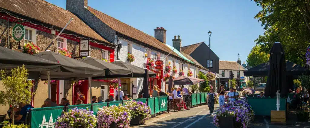 A charming street scene in a quaint village near Malahide Castle features people dining outdoors under umbrellas alongside colorful, flower-adorned buildings. The sky is clear, and the atmosphere is lively and inviting.
