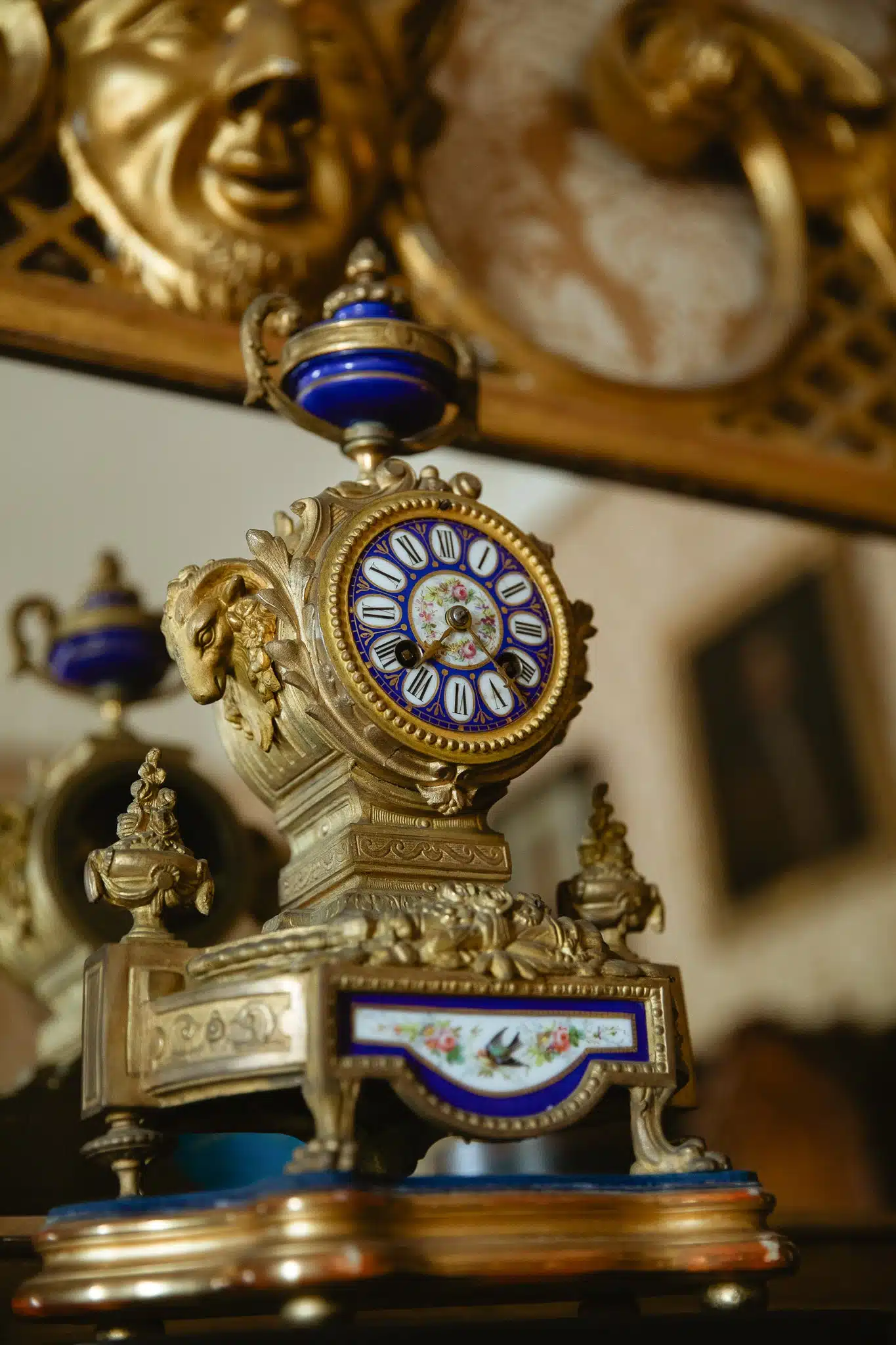 An ornate antique clock with an intricate gold frame and blue enamel detailing, reminiscent of the timeless elegance found in Malahide Castle. Roman numerals accentuate the clock face, which sits atop a decorative base set in a vintage interior with a blurred background.