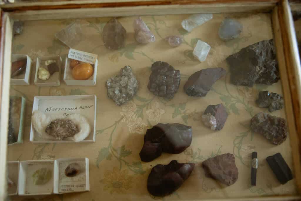 A display case at Malahide Castle contains a variety of minerals and crystals, including labeled and unlabeled specimens. The minerals range in color and texture, with some in small boxes and others placed directly on the floral-patterned surface.