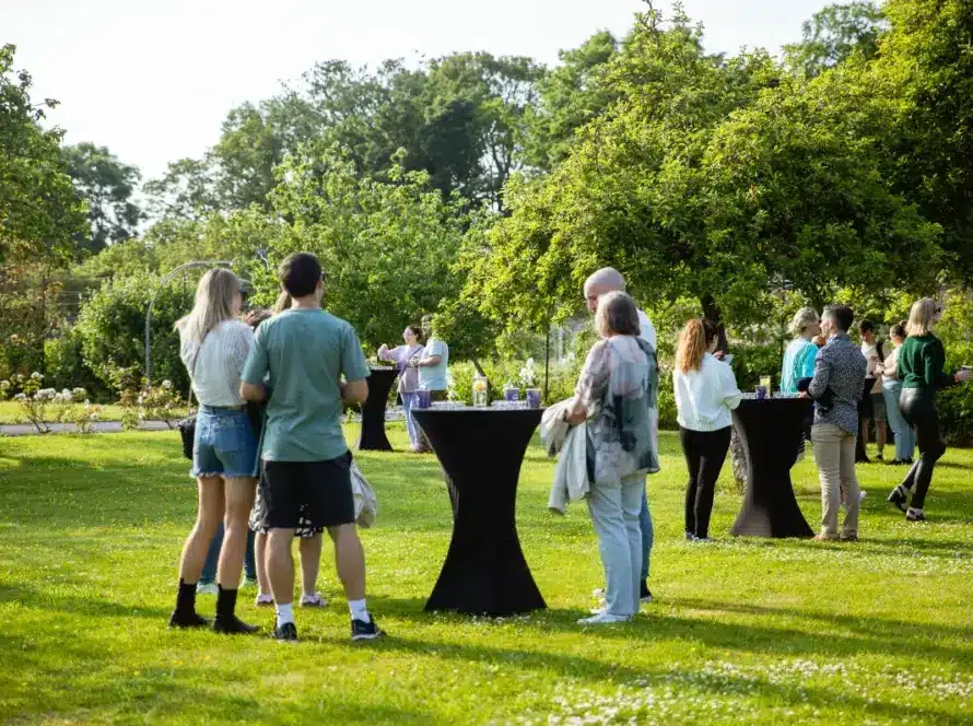 People are gathered in a grassy garden for a casual outdoor event at Malahide Castle. They stand in small groups around tall cocktail tables under sunny skies. Lush green trees surround the scene, giving it a vibrant, relaxed atmosphere.