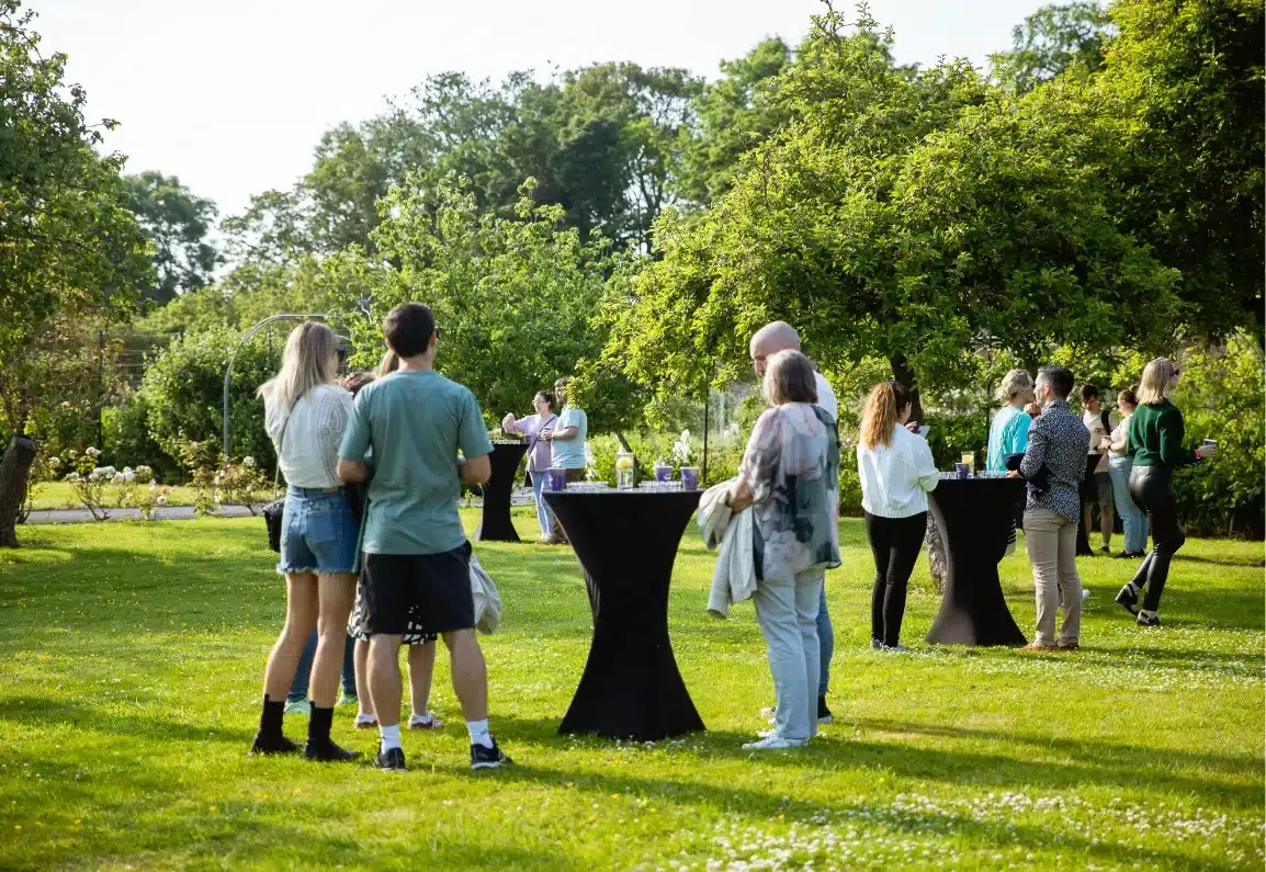 People are gathered in a grassy garden for a casual outdoor event at Malahide Castle. They stand in small groups around tall cocktail tables under sunny skies. Lush green trees surround the scene, giving it a vibrant, relaxed atmosphere.