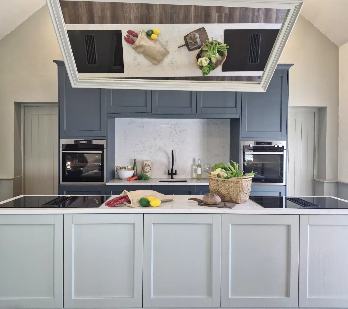 A modern kitchen with a light gray and navy color scheme. A marble countertop features a basket of greens, a knife, and vegetables. Two ovens are built into the cabinetry. The ceiling mirror reflects the countertop arrangement.