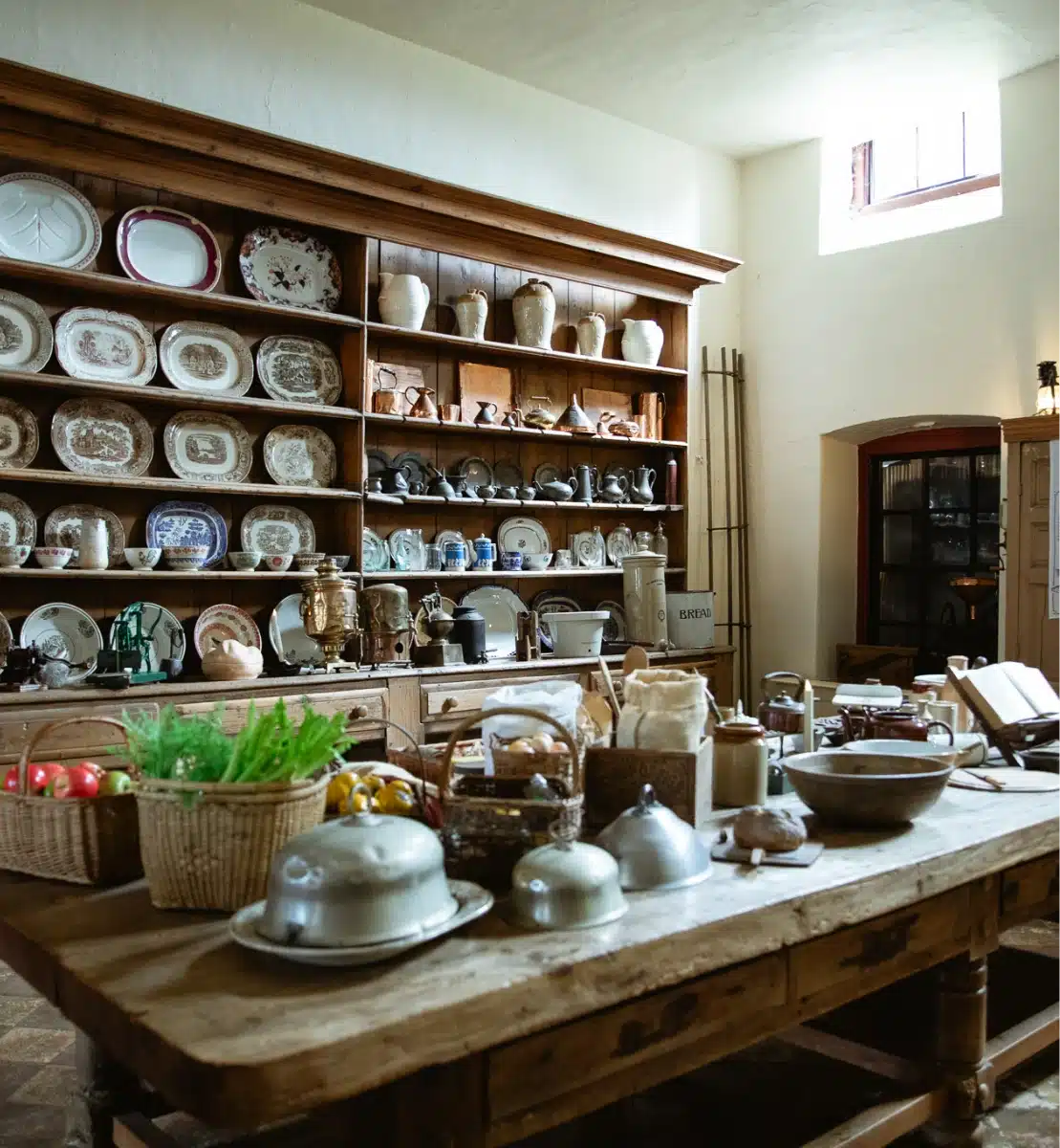 A rustic kitchen scene reminiscent of Malahide Castle, with wooden shelves displaying vintage plates, bowls, and jugs. A wooden table in the foreground holds baskets of produce, metal cloches, and kitchenware. Warm lighting from a small window completes the homely atmosphere.