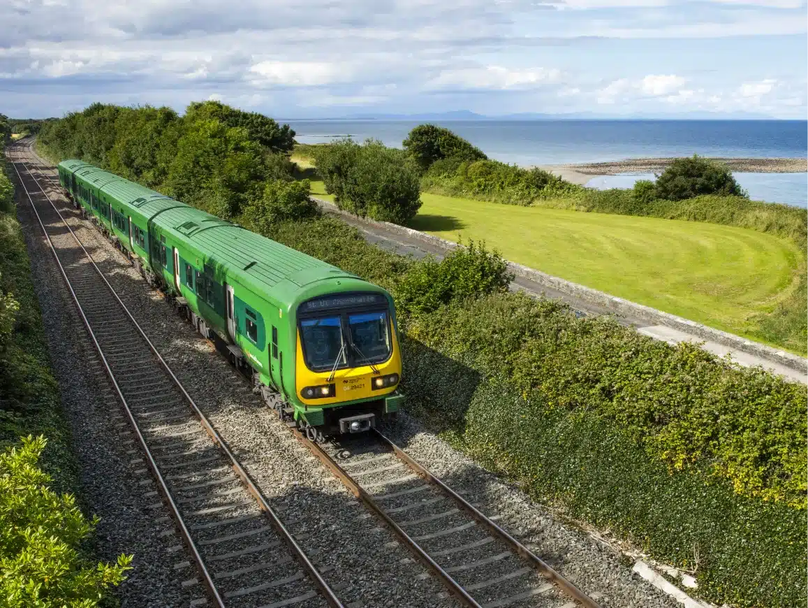 A green train travels along a railway with lush foliage on either side, near a scenic coastal area. The ocean and a grassy field are visible under a partly cloudy sky.