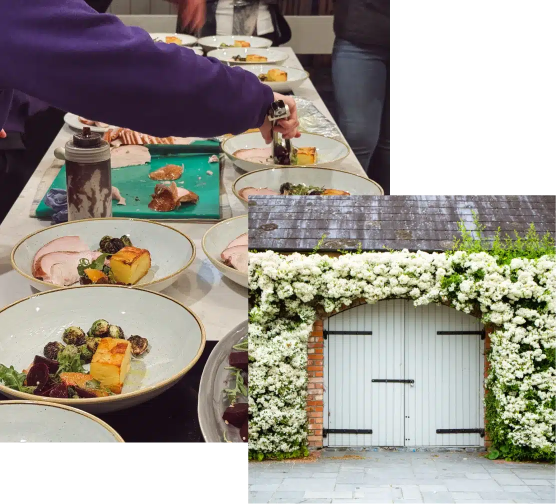A person prepares dishes with colorful vegetables and meat on a table. Next to it, a wooden gate is surrounded by lush white flowers on a brick wall.