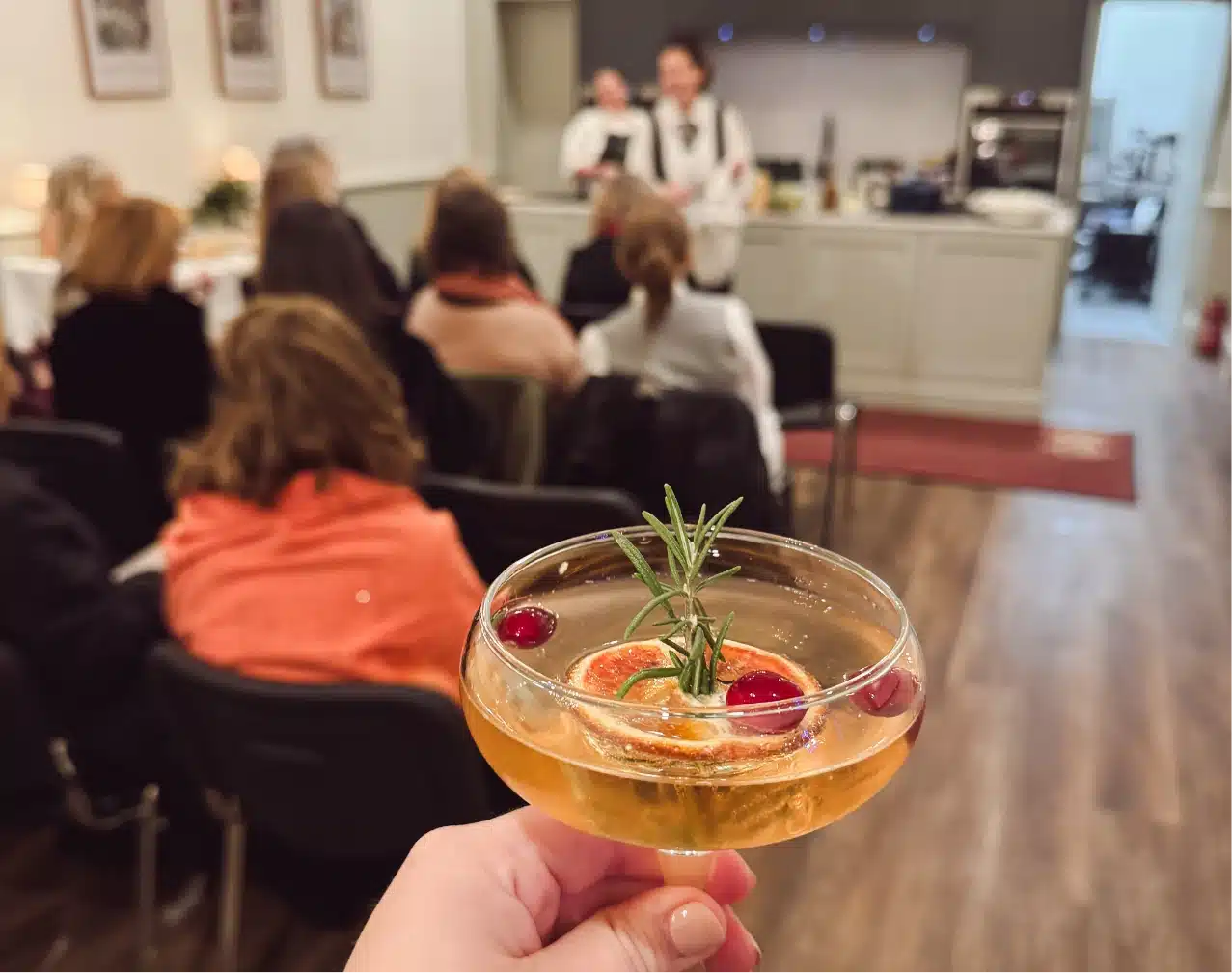 A hand holds a cocktail garnished with rosemary, a slice of orange, and berries. In the background, a seated audience listens to a presentation in a well-lit room with wooden flooring.